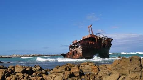 TimeLapse---Rusted-old-half-shipwreck-lying-next-to-rocky-coastline-of-L'Agulhas,-waves-running-in,-South-Africa