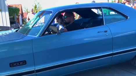 Blauer-Cadillac-Plymouth,-Der-Auf-Der-Straße-Läuft,-Während-Die-Leute-Im-Hintergrund-Zusehen
