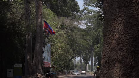 Weite-Außenaufnahme-Der-Straße-Und-Einer-Fliegenden-Kambodschanischen-Flagge,-Als-Ein-Mann-Hinter-Einem-Baum-Hervorkommt-Und-Mit-Einem-Schutzhelm-Auf-Die-Kamera-Zugeht