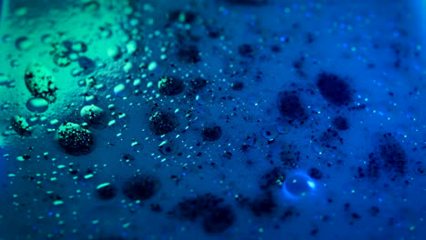 Liquid-with-oil-and-black-spots-on-surface-swirling,-lit-by-neon-colors,-Close-up