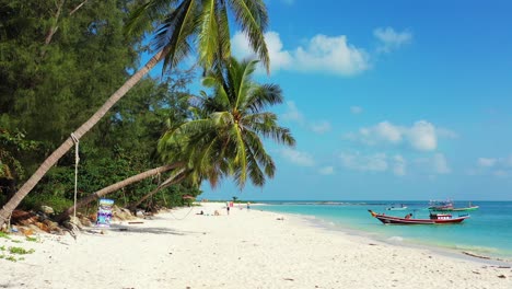 Peaceful-seascape-with-boat-floating-on-shore-of-tropical-island-with-white-sandy-beach-under-shadow-of-palm-trees-bent-over-in-Thailand