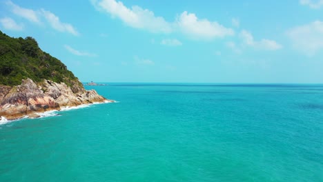 Rocky-slope-of-tropical-island-shoreline-splashed-by-sea-waves-on-a-bright-blue-sky-background-with-white-clouds-in-Thailand