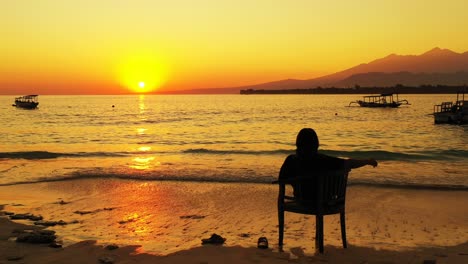 Silhouette-of-young-woman-sitting-on-a-chair-over-sea-flows,-enjoying-romantic-sunset-with-golden-colors-reflecting-on-calm-sea-surface-in-Bali