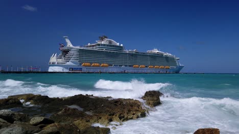 Cruise-ship-docked-at-Caribbean-Island-paradise-with-waves-breaking-on-a-rocky-shoreline-in-foreground