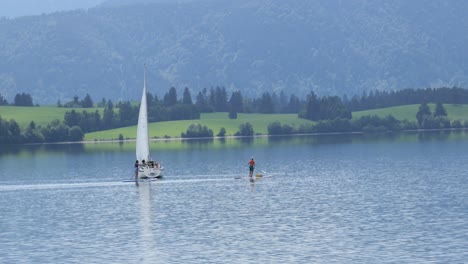 Sailing-Boat-and-Stand-Up-Paddlers-on-Lake-Forggensee-near-Fuessen-in-Bavaria,-Germany
