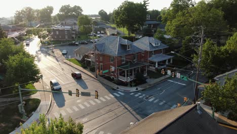 Static-aerial-establishing-shot-of-small-town-in-America,-house-on-corner-of-intersection-at-traffic-signal-light,-cars-stopped-at-light,-traffic-drives-on-road,-drone-above-rooftop,-summer-light