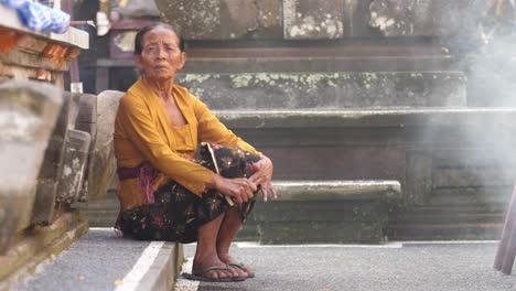 Elderly-woman-sits-on-stone-step-in-Hindu-temple-on-island-of-Bali,-Indonesia-with-smoke-from-fire-and-incense-fills-scene