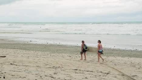 Rückaufnahme-In-Zeitlupe-Zwei-Junge-Surferinnen,-Die-Ein-Surfbrett-Halten,-Gehen-In-Tambor,-Costa-Rica,-Am-Strand-Entlang