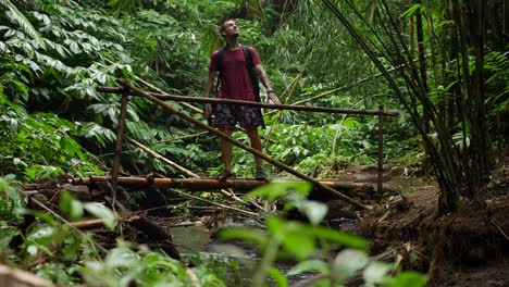 A-young-man-in-t-shirt-and-shorts-walks-over-a-small-bamboo-bridge-over-a-small-stream-of-water,-looks-at-the-beautiful-surroundings-and-continue-crossing