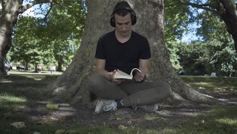 Young-white-man-seated-under-a-tree-reading-a-book-in-a-park-in-Cambridge-city,-England