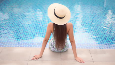 Sexy-woman-sitting-on-the-edge-of-the-swimming-pool-at-an-exotic-hotel-in-Florida-wearing-botched-spotty-monokini-and-white-hat,-back-view-slow-motion-handheld