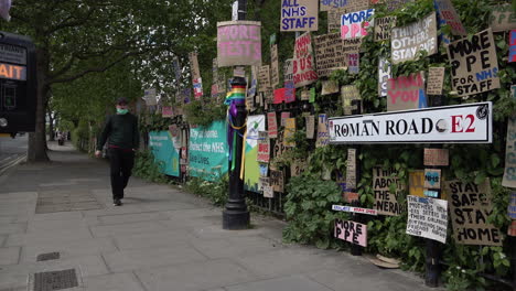 A-man-in-a-face-mask-walks-past-a-huge-memorial-wall-of-cardboard-signs-showing-support-of-healthcare-staff-during-the-Coronavirus-outbreak-line-the-edge-of-a-park-in-the-East-End-of-London