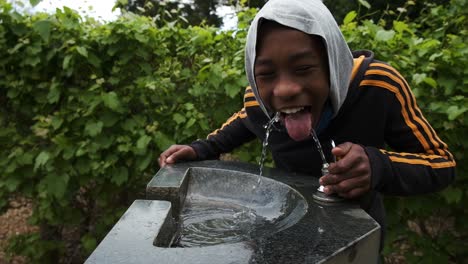 Happy-young-black-male-drinking-fresh-water-from-outdoor-fountain-faucet