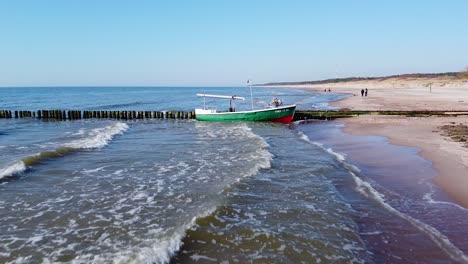 Beautiful-aerial-view-of-a-red-tracked-tractor-and-green-coastal-fisherman-fishing-boat-docked-at-the-old-wooden-pier-in-sunny-day,-calm-sea-shore,-wide-angle-low-altitude-drone-shot-moving-forward
