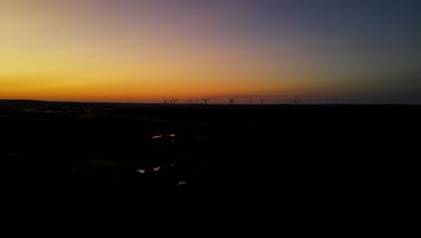 Luftaufnahme---Wegfliegen-Vom-Windpark-Bei-Sonnenuntergang,-Windturbinensilhouette-Weit-Entfernt-Im-Hintergrund,-Puck,-Pomorskie,-Polen