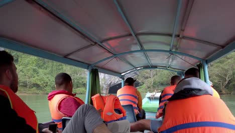 Tour-boat-with-people-in-life-jackets-approaching-cavern-entrance-beneath-the-mountain,-Handheld-shot
