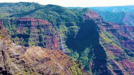 HD-Hawaii-Kauai-slow-motion-static-wide-shot-of-Waimea-Canyon-with-a-helicopter-entering-frame-right-flying-toward-a-waterfall-in-the-distance