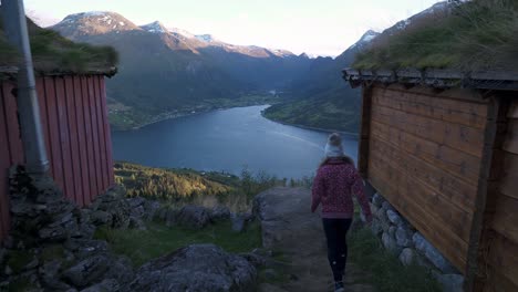 Girl-walking-in-between-two-small-and-old-cabins,-towards-view-point,-watching-over-a-beautiful-scenery-of-mountains-and-a-large-lake-in-Loen,-Norway