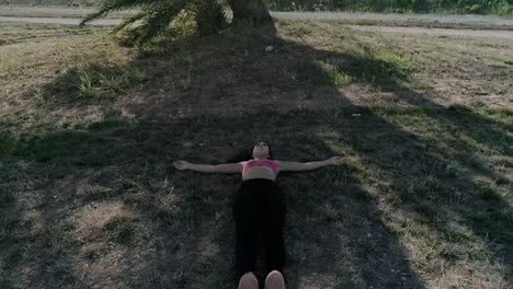 Drone-shot-of-Young-Brunette-Woman-lying-on-the-ground-next-to-a-big-tree