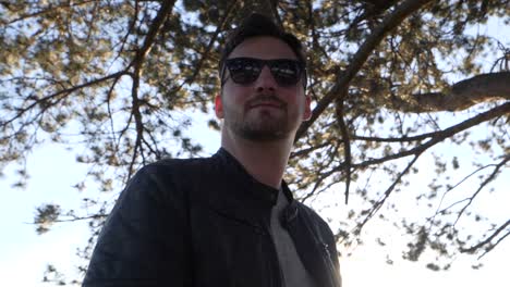 Young-bearded-man-with-sunglasses-and-leather-jacket-stands-in-sunshine-under-a-tree