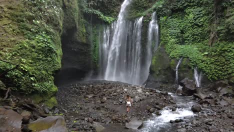 Woman-walking-over-rocks-towards-giant-waterfall-in-rainforest