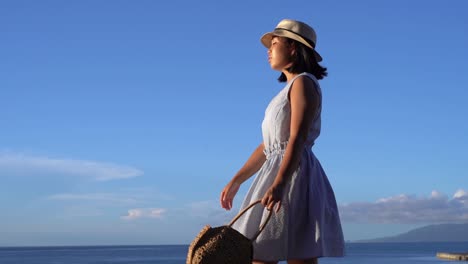 Japanese-girl-in-summer-dress-holding-bag-walking-in-front-of-Ocean-on-beautiful-clear-day---sideways-tracking-shot-SLOW-MOTION