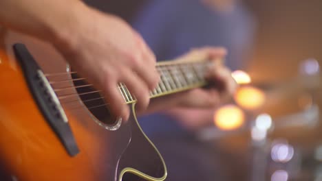 Close-up-of-a-professional-musician-playing-western-guitar-with-a-blurred-drummer-in-the-background-during-a-live-session