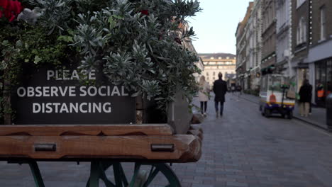 People-walk-past-a-sign-on-a-decorative-handcart-that-says,-“Please-observe-social-distancing”-in-a-very-quiet-Covent-Garden-during-the-second-national-Coronavirus-lockdown