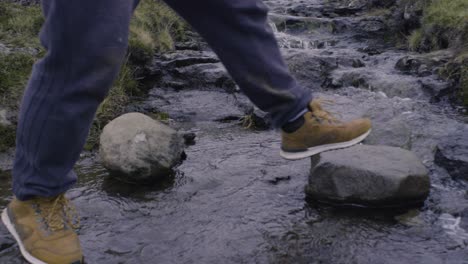 Close-up-of-feet-walking-across-a-small-river-creek-in-brown-hiking-boots-and-dirty-pants-in-nordic-landscape-in-slow-motion-on-The-Faroe-Islands