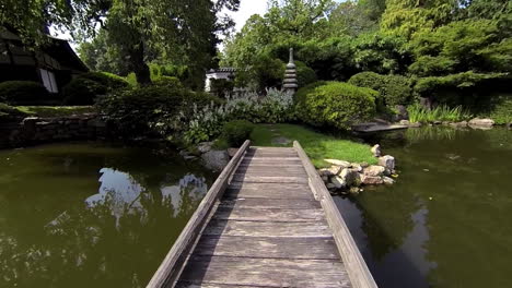 Steadicam-shot-crosses-foot-bridge-of-pond-up-to-stone-pagoda,-flowers-and-Japanese-lantern