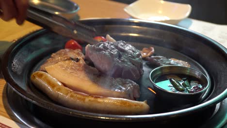 Person-Grilling-Sausage,-Pork-Belly,-And-Tomatoes-On-Griller-In-Korean-Restaurant