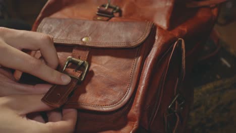 Woman's-Hands-Opening-The-Pocket-Belt-Buckle-Of-A-Leather-Retro-Brown-Backpack---Closeup-Shot