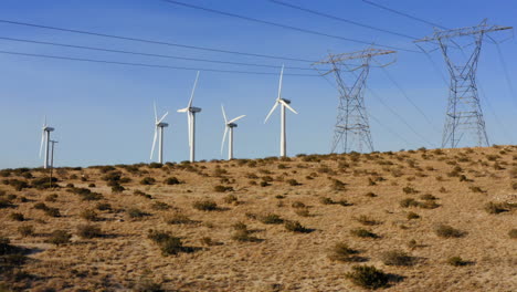 Vista-Aérea-Panorámica-De-Derecha-A-Izquierda-Con-Vistas-A-Las-Turbinas-Eólicas-Y-La-Línea-De-Transmisión-De-Alto-Voltaje-Que-Revela-Un-Enorme-Parque-Eólico-Cerca-De-Palm-Springs-En-El-Desierto-De-Mojave,-California,-EE.UU.