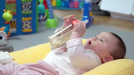 Healthy-Baby-Girl-Lying-On-Back-In-Cushion-Playing-Her-Feeding-Bottle