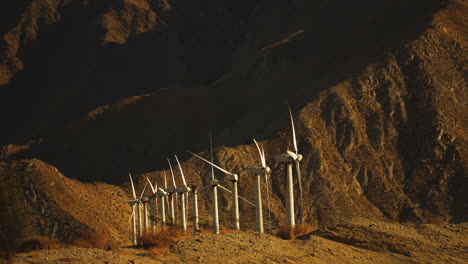 Miniature-looking-wind-turbines-rotating-at-wind-farm-in-the-desert-with-huge-mountain-in-the-background-near-Palm-Springs-in-the-Mojave-Desert,-California,-USA