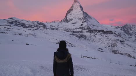 Girl-walking-infront-of-Famous-Matterhorn-Mountain-at-sunrise,-red-dawn-sky-winter-wonderland-at-swiss-alps-zermatt-glacier-ski-resort