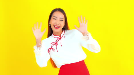 Pretty-Happy-Asian-Girl-Waving-at-Camera-in-front-of-yellow-Backdrop