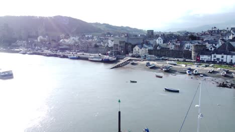 Idyllic-Conwy-castle-and-harbour-fishing-town-boats-on-coastal-waterfront-aerial-pull-away-fast