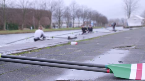 Panning-shot-with-female-rowers-preparing-for-a-training-session-in-single-scull-and-double-scull-boats,-with-green-white-and-red-oar-in-the-foreground-Sport-fitness