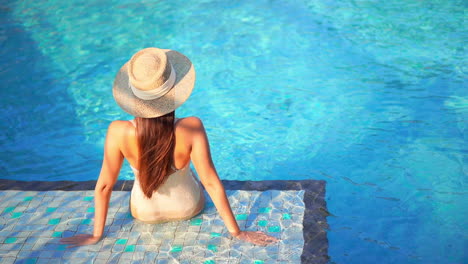 Back-of-attractive-female-sitting-on-edge-of-blue-swimming-pool-on-sunny-summer-day,-full-frame