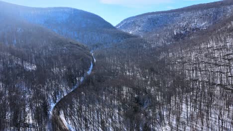Aerial-drone-footage-of-a-snowy-mountain-valley-and-road-in-early-spring-on-a-sunny-day-in-the-Appalachian-Mountain-Range,-just-after-winter-ends-with-forests-and-snow-and-sunshine-and-blue-skies