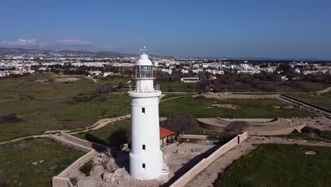 Stunning-rotating-drone-footage-circling-a-white-lighthouse-in-Cyprus,-Greece-with-the-city-in-the-background