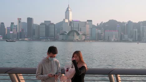 A-couple-takes-a-photo-while-holding-a-boutique-of-roses-along-the-Victoria-Harbour-waterfront-and-Hong-Kong-Island-skyline-in-the-background-while-the-sunset-sets-in