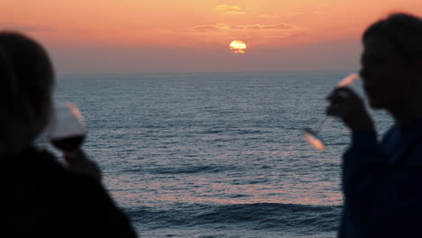 Silhouette-Von-Zwei-Mädchen,-Die-Einen-Rotwein-Vor-Dem-Wunderschönen-Meer-Und-Dem-Goldenen-Sonnenuntergang-Im-Hintergrund-Trinken