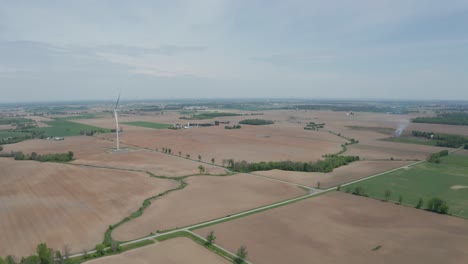 Wide-aerial-view-of-flat-terrain,-farm-fields-and-large-wind-power-turbine-towering-over-the-landscape