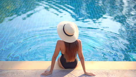 Unrecognizable-Woman-with-Sun-Hat-Sitting-by-the-Pool,-High-Angle-Rear