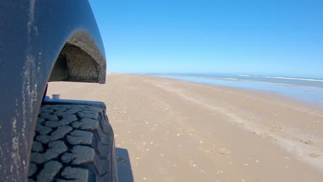 Right-front-tire-of-vehicle-and-beach-surf-driving-on-the-sand-on-a-sunny-day-on-South-Padre-Island-Texas--Point-of-view,-POV