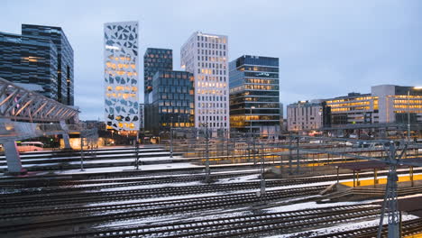 Tren-Que-Sale-De-La-Estación-Central-De-Oslo-Con-Edificios-De-Proyecto-De-Código-De-Barras-Por-La-Noche-En-Bjorvika,-Oslo,-Noruega
