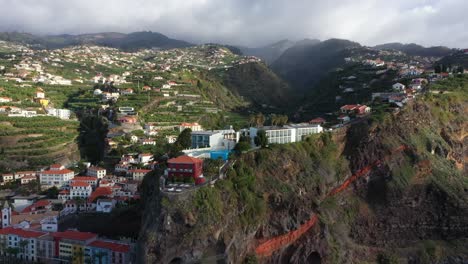 Circling-aerial-shot-of-luxury-villas-and-a-hotel-on-top-of-a-cliff-in-Madeira-with-a-village-in-the-background-and-dark-clouds-on-top-of-the-mountain