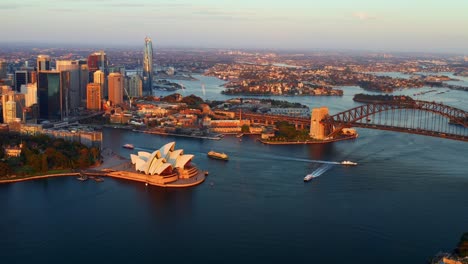 Zoom-out-Aerial-View-of-Sydney-Harbour-Bridge-and-Opera-House-with-Ferry-Boats-Cruising-at-Sunset
